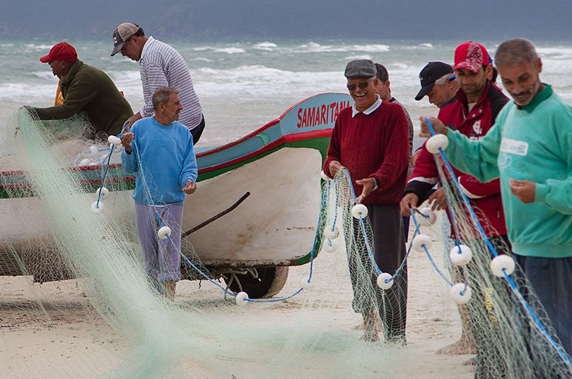 Pescadores trabalham com rede e embarcação na praia.
